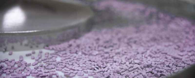A stainless steel machine with a conveyor belt moves hundreds of purple pills along a Sanofi manufacturing line.