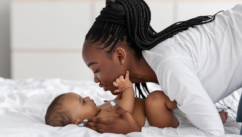 A mother with braided hair and a white long-sleeved shirt lies on a white bed, leaning close to her infant. The baby lies on the bed partially covered by white sheets, wearing a diaper, with one hand gently resting on the mother's chest. The baby's fingernails are painted blue. The setting is a cozy bedroom, and the image conveys a warm, intimate bond between mother and child.