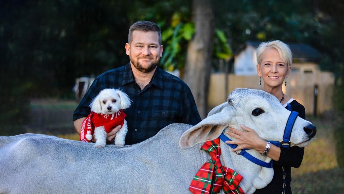 Man and woman smiling, holding a dog and posing with a cow