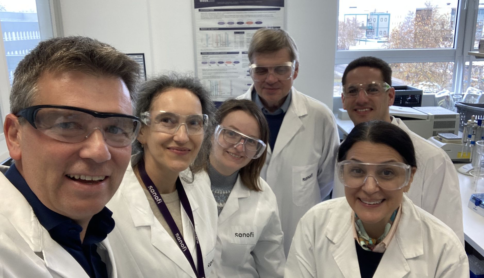 Six Sanofi employees are wearing white lab coats in a lab that has windows overlooking Frankfurt, Germany