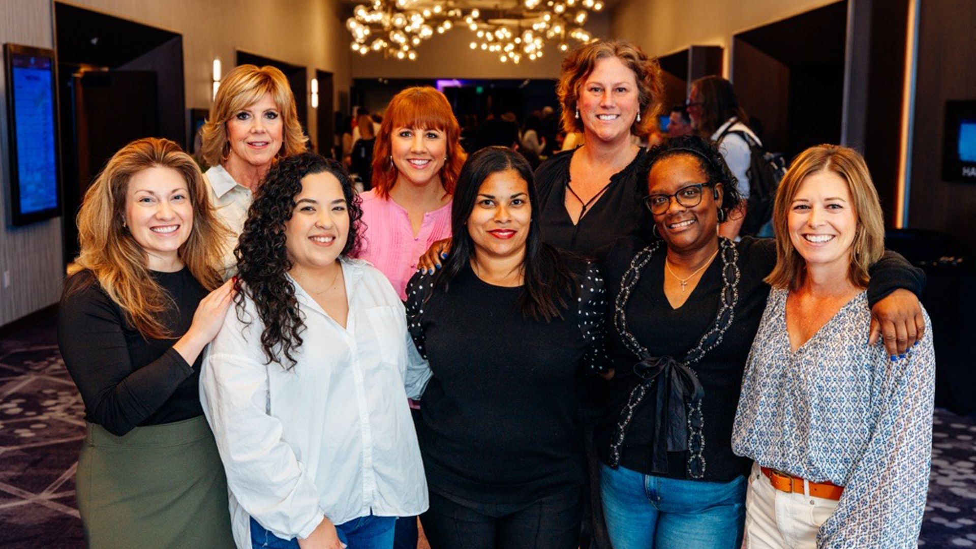 Eight women pose in business casual attire in a bright conference space