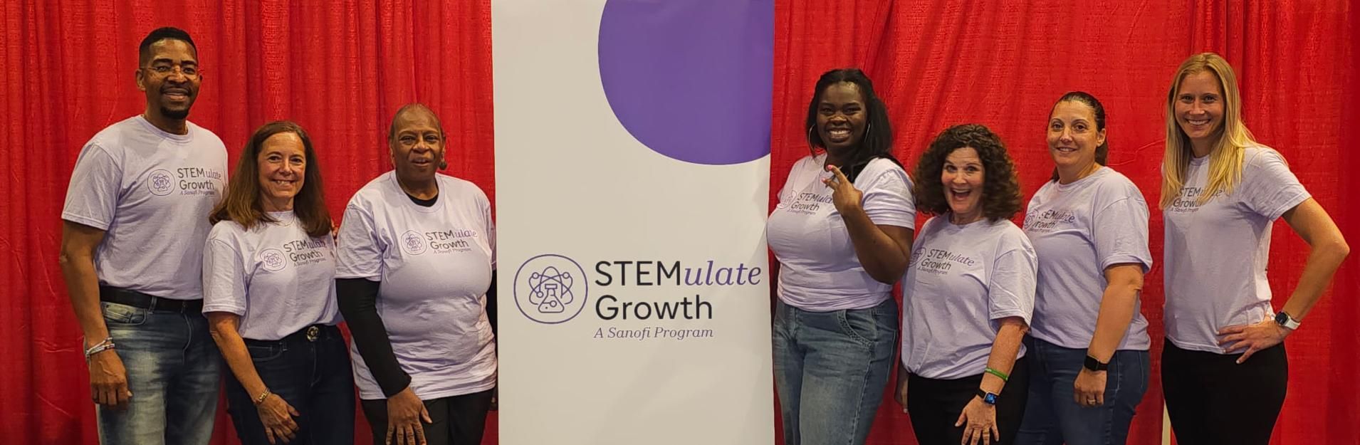 Seven Sanofi team members in matching purple “STEMulate Growth” t-shirts stand smiling beside a program banner with a red curtain backdrop.