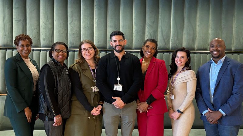 Seven smiling Sanofi SCAN team members in business professional attire standing together for a group portrait against a green textured background.