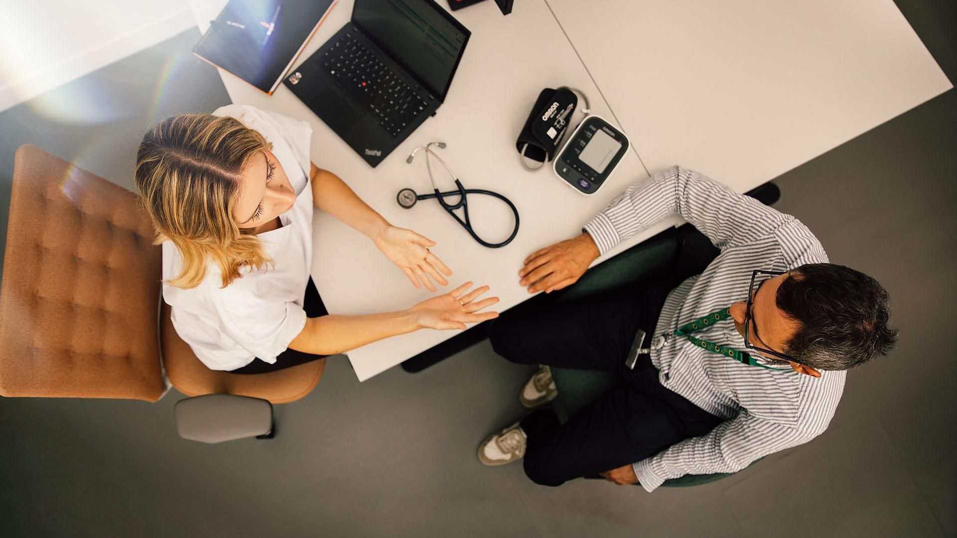 Overhead view of a desk with a healthcare professional and a patient in conversation, stethoscope and laptop visible.