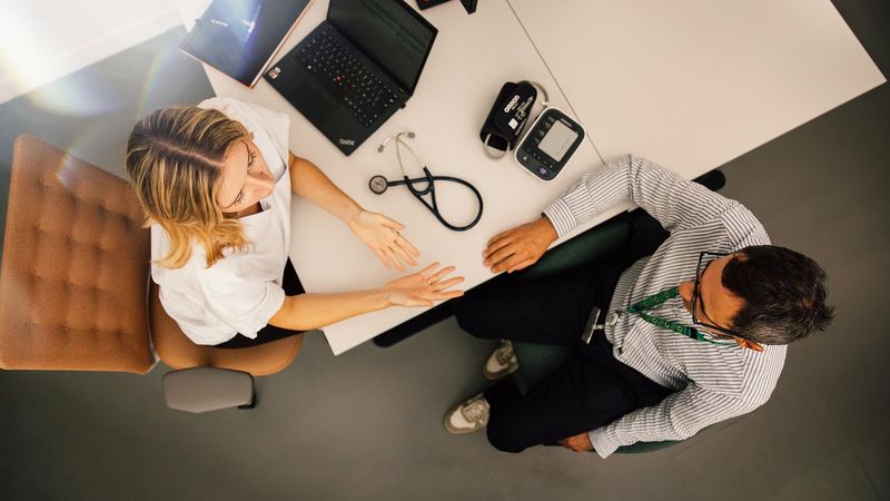 Overhead view of a desk with a healthcare professional and a patient in conversation, stethoscope and laptop visible.