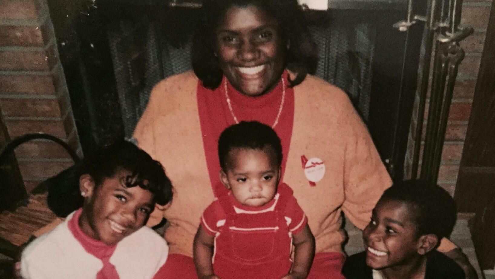 Vintage family photograph from the 1980s featuring a smiling woman seated with three children in holiday sweaters, taken in front of a brick fireplace in a home setting