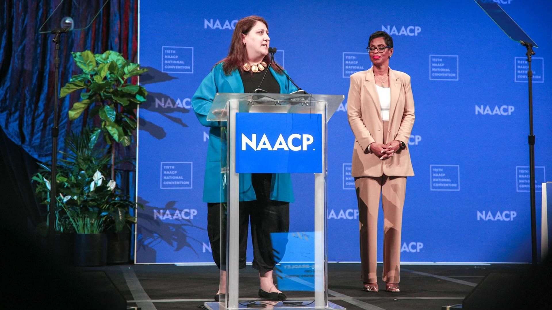 On stage at the 115th NAACP National Convention, Deb Glasser of Sanofi, wearing a bright blue blazer and black outfit, announces a collaboration with the NAACP from a podium. Karen Boykin-Towns of NAACP, in a beige suit and glasses, stands beside her with hands clasped.