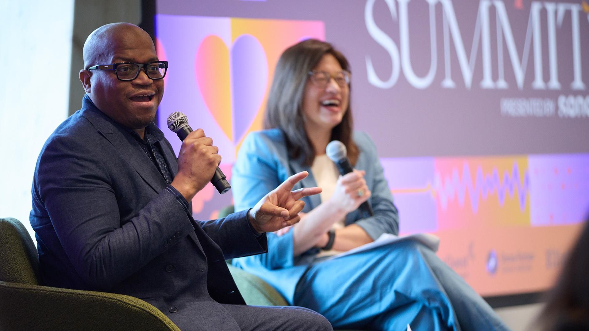 Jamal Watkins, in a dark suit and glasses, gestures while speaking into a microphone during a panel discussion at the Boston Globe Health Equity Summit. Seated beside him, a panelist in a light blue blazer listens and smiles in front of a colorful "Summit" backdrop.