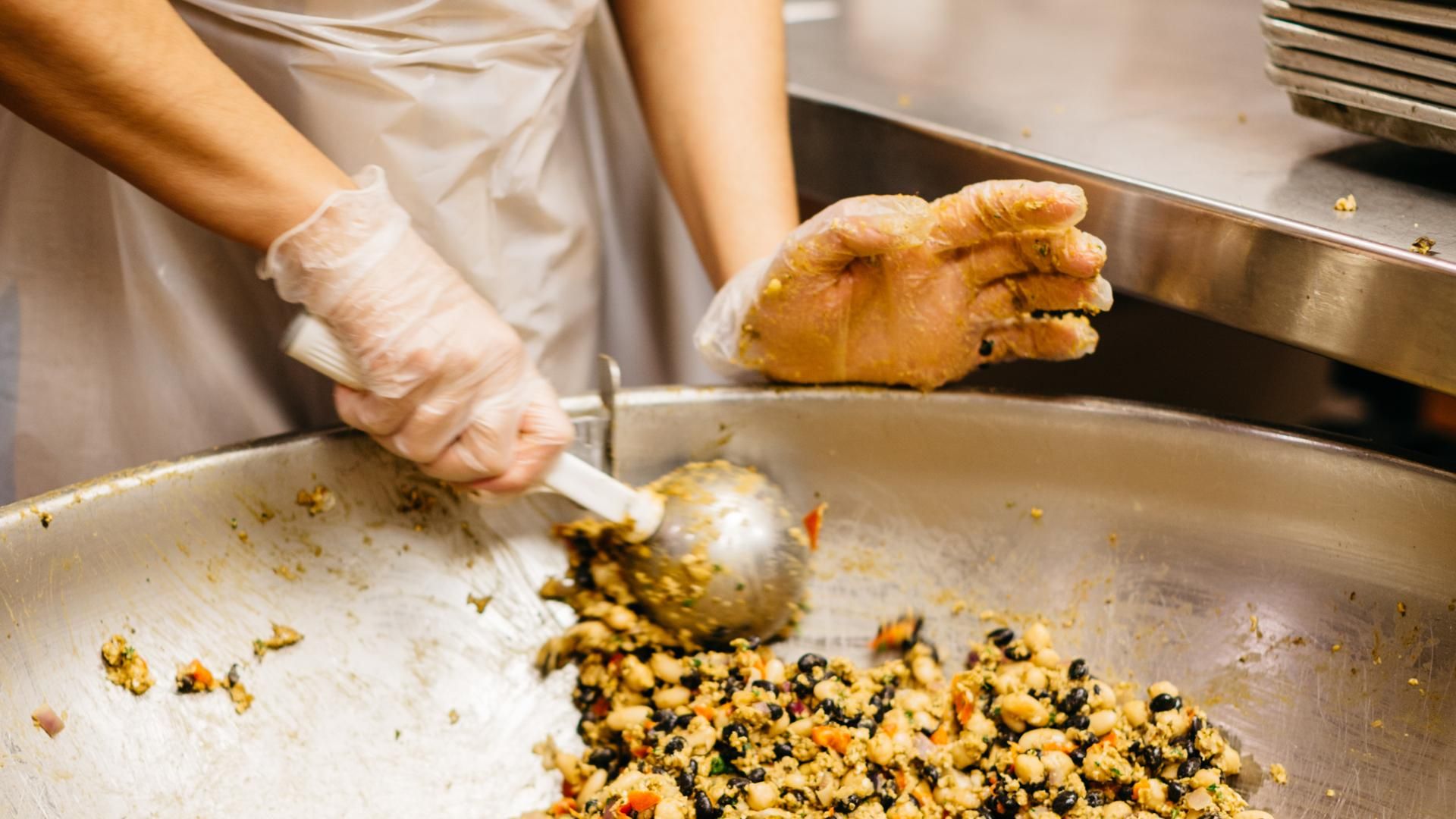 A food service worker wearing gloves prepares a bean and vegetable dish in a commercial kitchen.