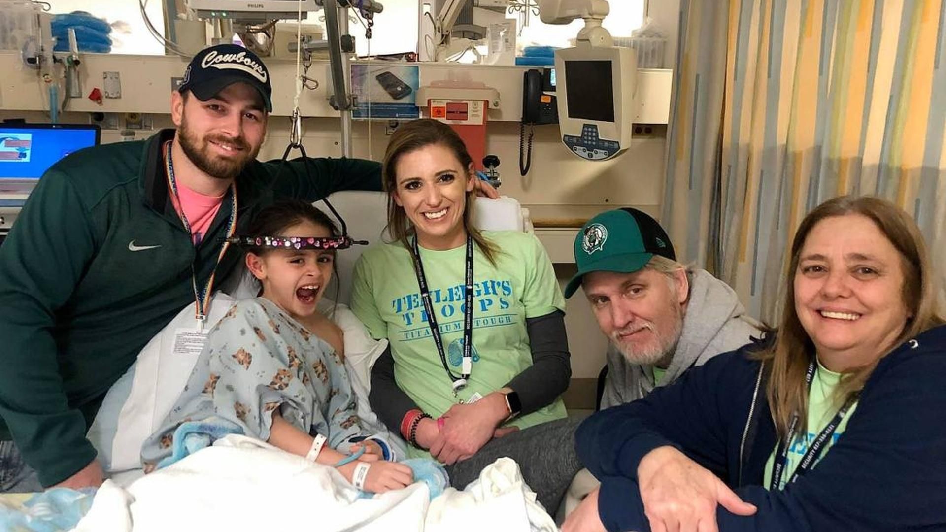 A young patient in a hospital bed smiles alongside four adults in a hospital room.