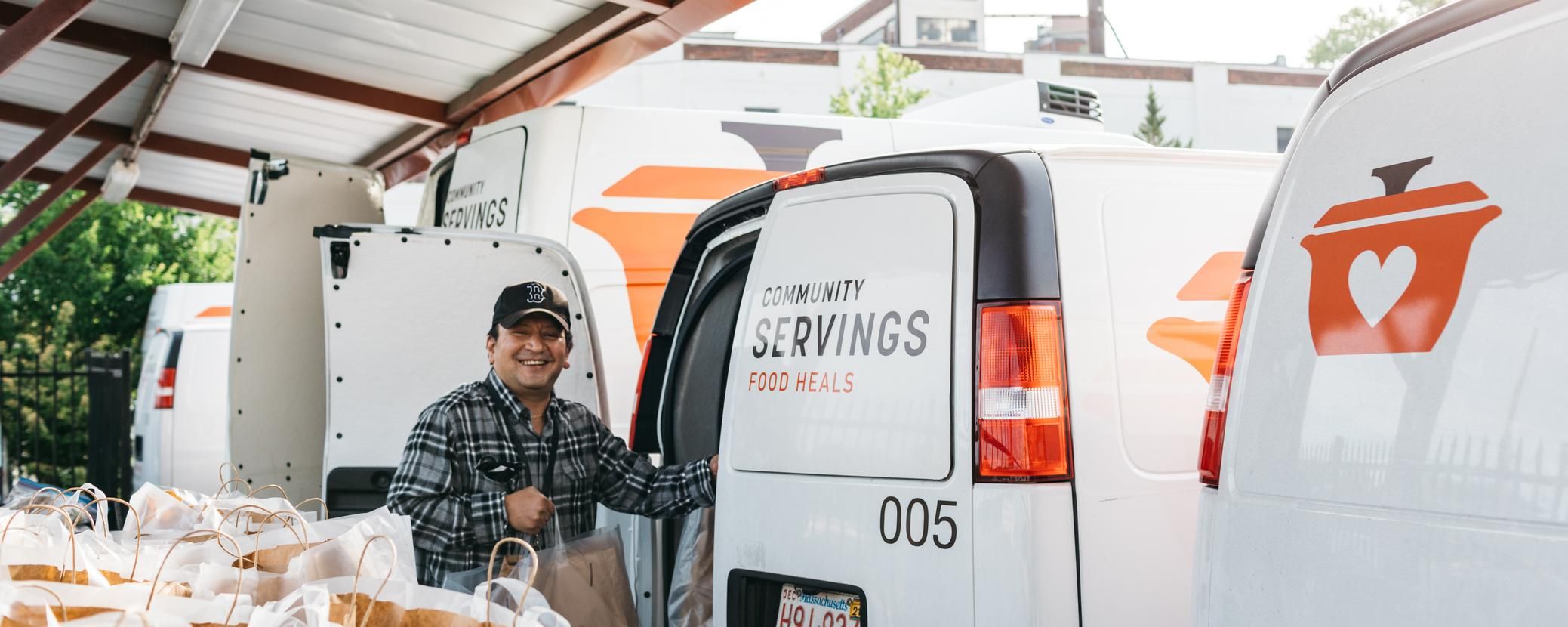 A Community Servings team member stands beside a branded delivery van with bags of prepared meals ready for delivery.