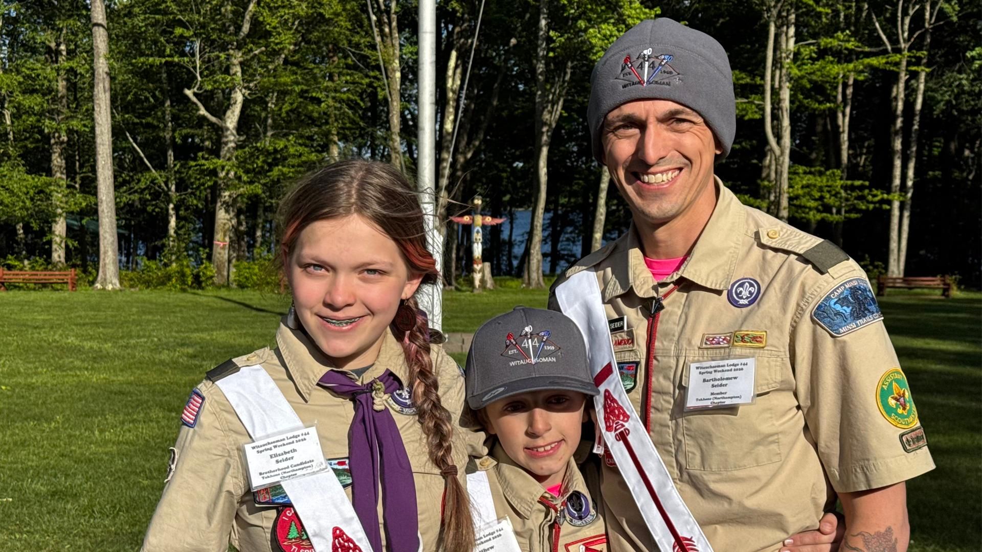 Bart Seider stands with his two children in their Scouting uniforms on a grassy outdoor field; his daughter wears a Scout uniform with a purple neckerchief, and his son wears a Scout uniform with a red bola.