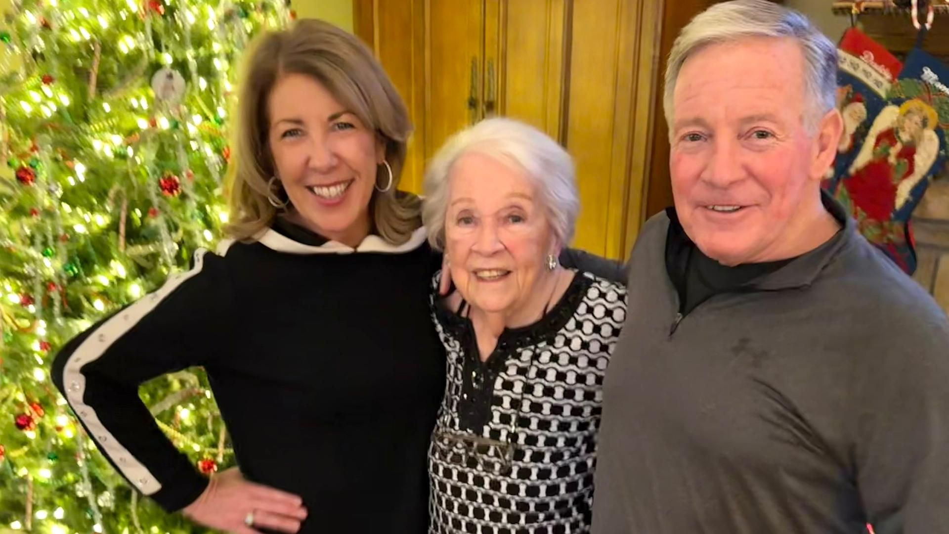 Three people smiling and posing together in front of a decorated Christmas tree. On the left is Mary Dugan, a smiling woman with short light brown hair, wearing a black top with white sleeves and a ring on her left hand. In the middle is her mother, a woman with short white hair, wearing a black and white patterned top. On the right is her brother Jerry, a man with white hair, wearing a gray shirt. The tree has white lights, red and green ornaments, tinsel, and Christmas stockings hang on the wall behind them.