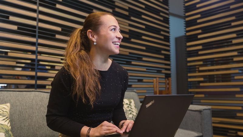 A woman with long, wavy brown hair sits on a gray couch, smiling while looking at a ThinkPad laptop on her lap. She wears a black long-sleeve top and earrings, with a wooden slatted wall in the background.
