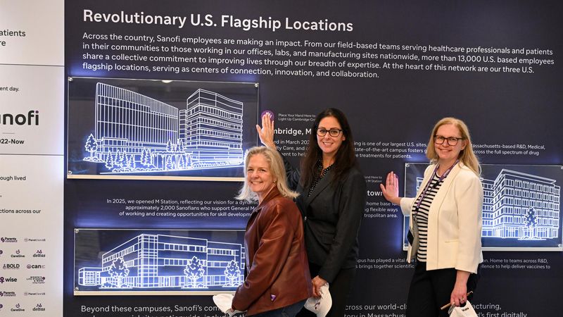 Three Sanofi employees pose in front of a large banner titled "Revolutionary U.S. Flagship Locations" featuring images and information about Sanofi's key U.S. campuses including M Station in Morristown, during the America 250 immersive employee engagement experience