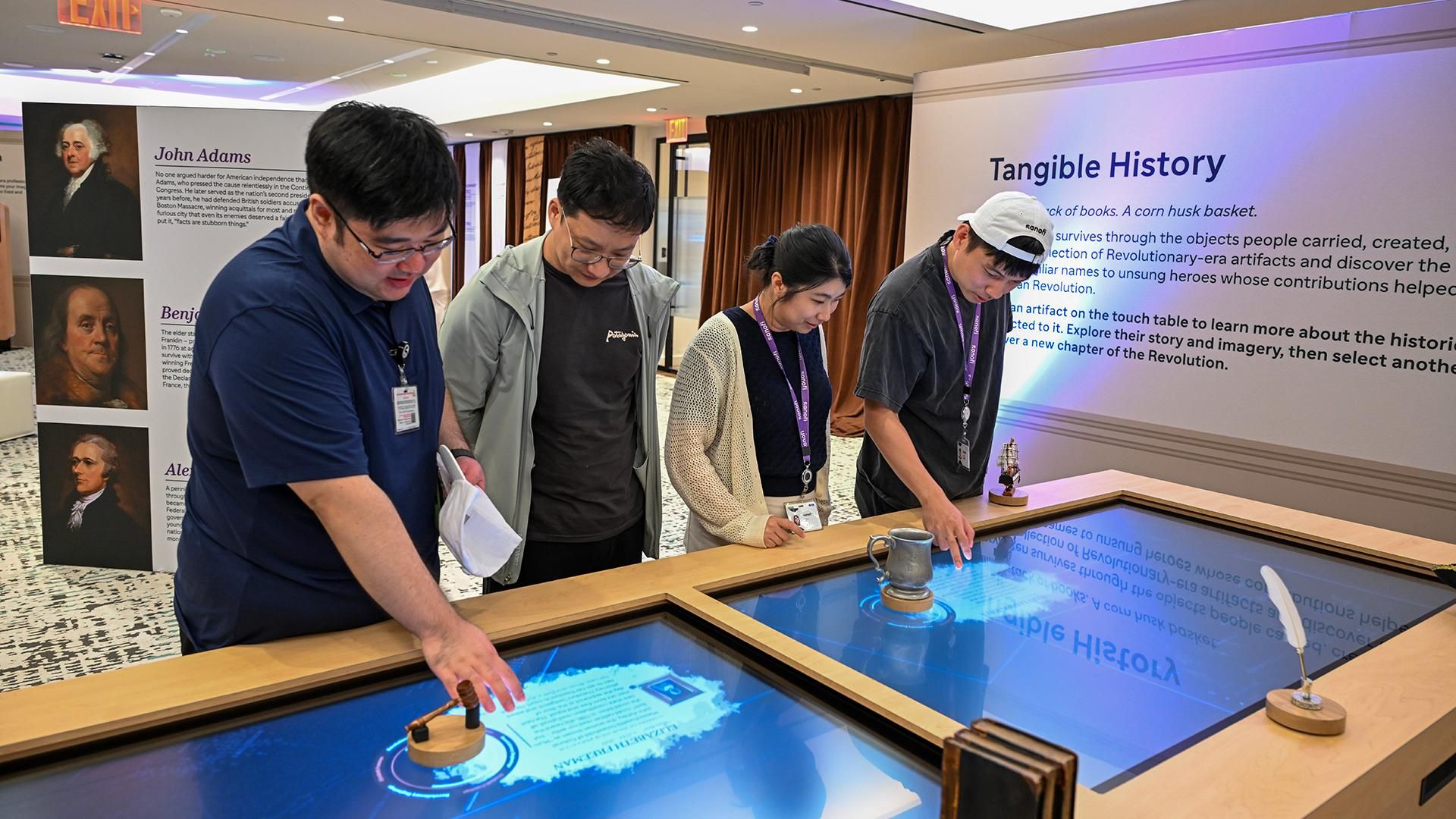 Four Sanofi employees interact with a digital touch table displaying an interactive map and 3D rotating models of historical artifacts, including a quill pen and corn husk basket, at the America 250 "Tangible History" immersive exhibit.