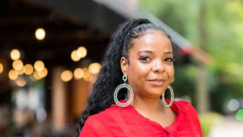 Anise, an aTTP patient in the US, wears a red shirt and stands outside in a forested area against a pavilion lit by white lights.