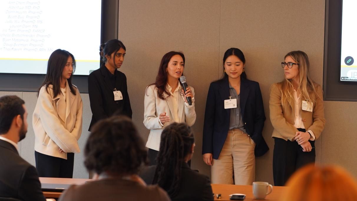Five UCSF School of Pharmacy students present their winning immunology go-to-market strategy to a seated panel at the IPhO VIP Case Competition, with judges' names displayed on the screen behind them.