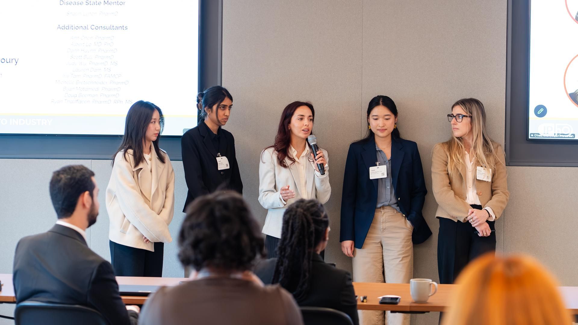 Five UCSF School of Pharmacy students present their winning immunology go-to-market strategy to a seated panel at the IPhO VIP Case Competition, with judges' names displayed on the screen behind them.