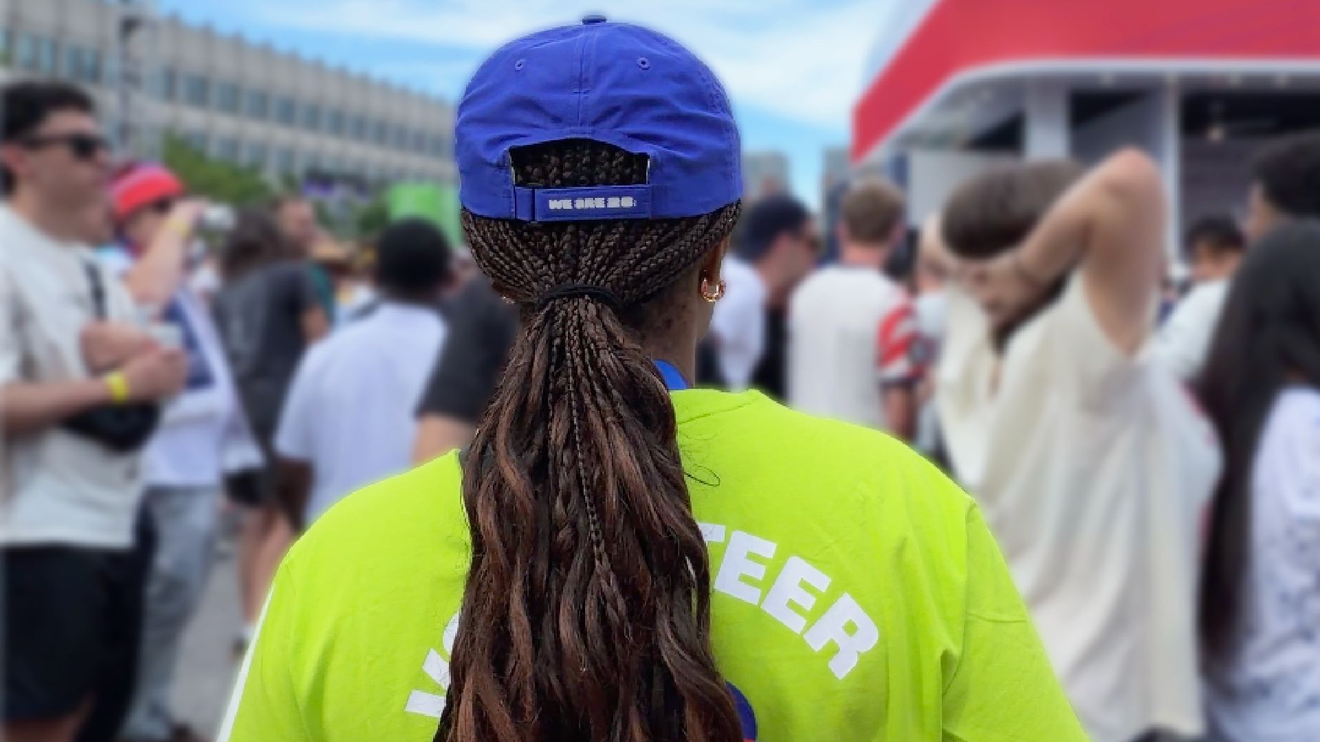 Gigi is standing wearing a bright volunteer shirt and blue cap with her back to the camera overlooking a crowded outdoor fan event, with people, tents, and city buildings in the background at a World Cup Boston Fan Fest.