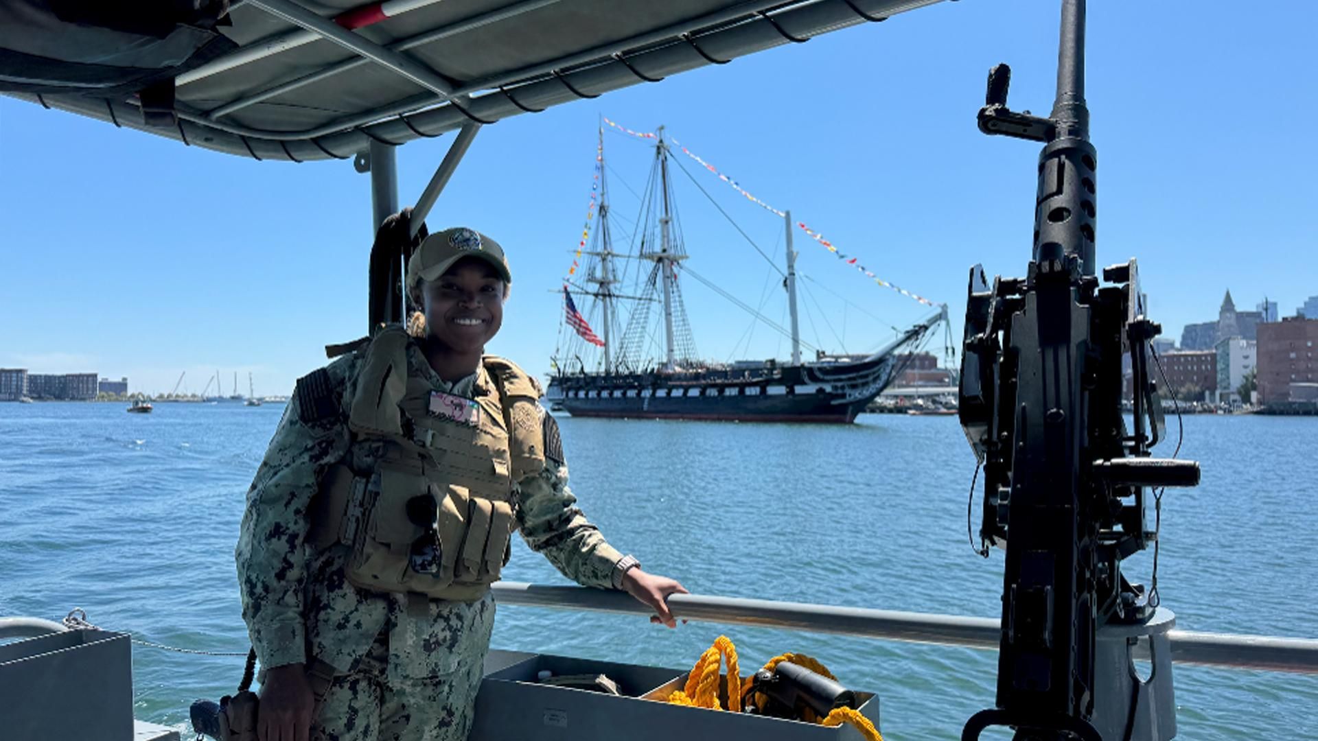 Sanofi Senior Associate Scientist Gigi Abellard is seen in a camouflage uniform and life vest standing on a naval vessel holding a railing. Onboard equipment is seen in the foreground and the USS Constitution sailing ship with masts and American flag visible in the background under a clear blue sky.