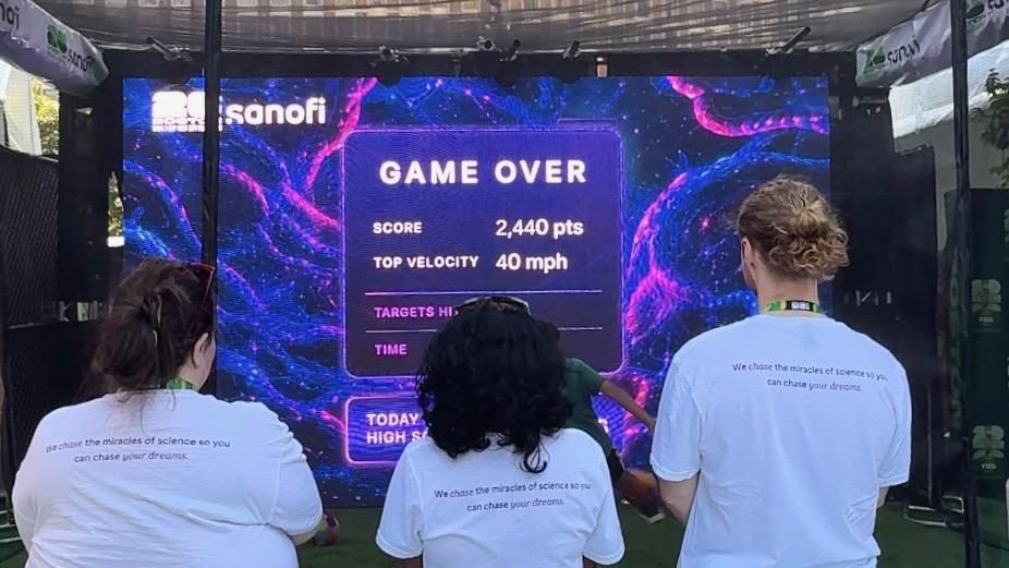 Three participants are wearing white branded t-shirts Sanofi t-shirts that state “We chase the miracles of science so you can chase your dreams.” All participants stand facing a large interactive digital screen at a Boston Fan Fest immersive experience. The screen displays a “Game Over” results page with a score of 2,440 points and a top velocity of 40 mph, set against a vibrant, virus-themed graphic background in blue and purple. The activation uses motion-sensing technology to simulate a sports challenge where players strike virtual virus targets. The display includes Sanofi branding, and event surroundings with structural supports and banners are visible around the installation.