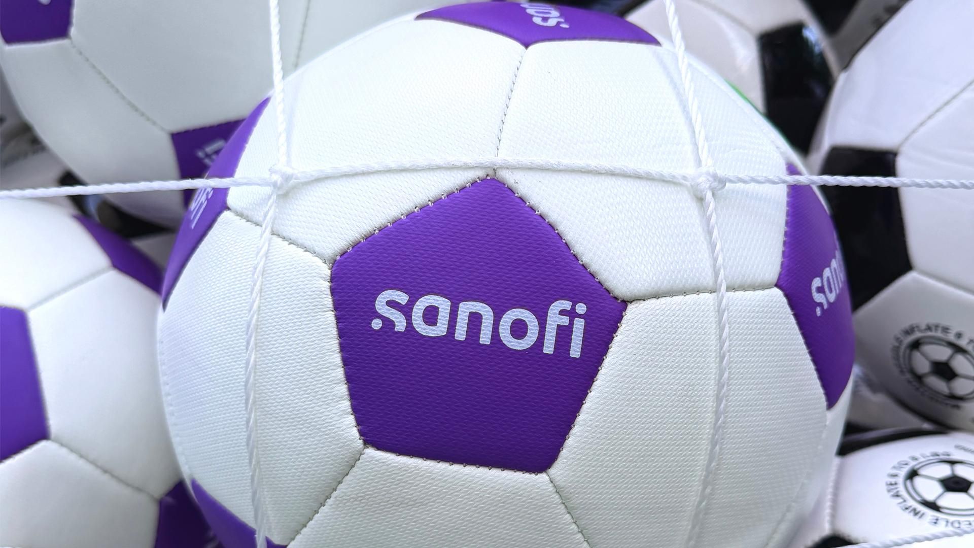 Dozens of soccer balls are held in a net display, some in the standard black and white and others in Sanofi miracle purple and white; the Sanofi logos is visible