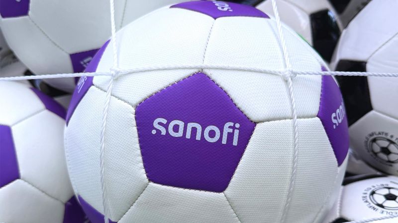 Dozens of soccer balls are held in a net display, some in the standard black and white and others in Sanofi miracle purple and white; the Sanofi logos is visible