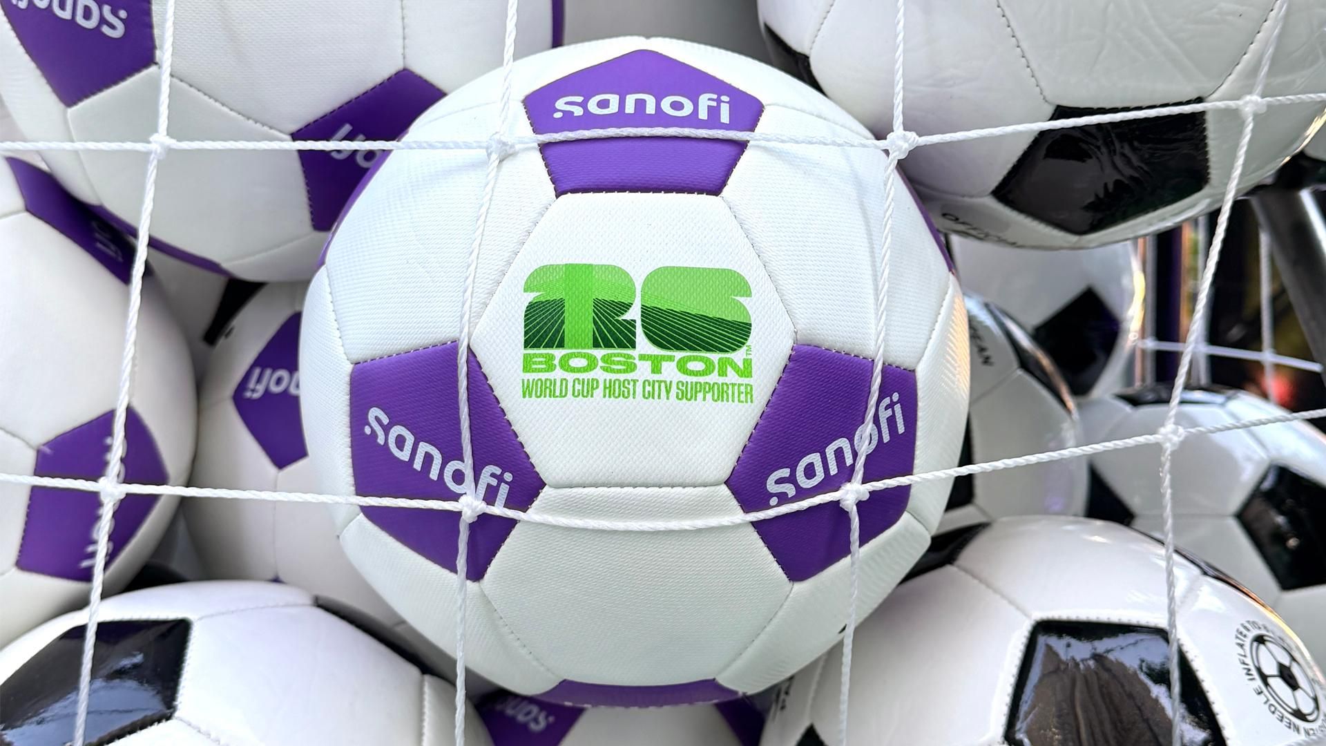 Dozens of soccer balls are held in a net display, some in the standard black and white and others in Sanofi miracle purple and white; both the Sanofi and Boston World Cup Host City Supporter logos are visible
