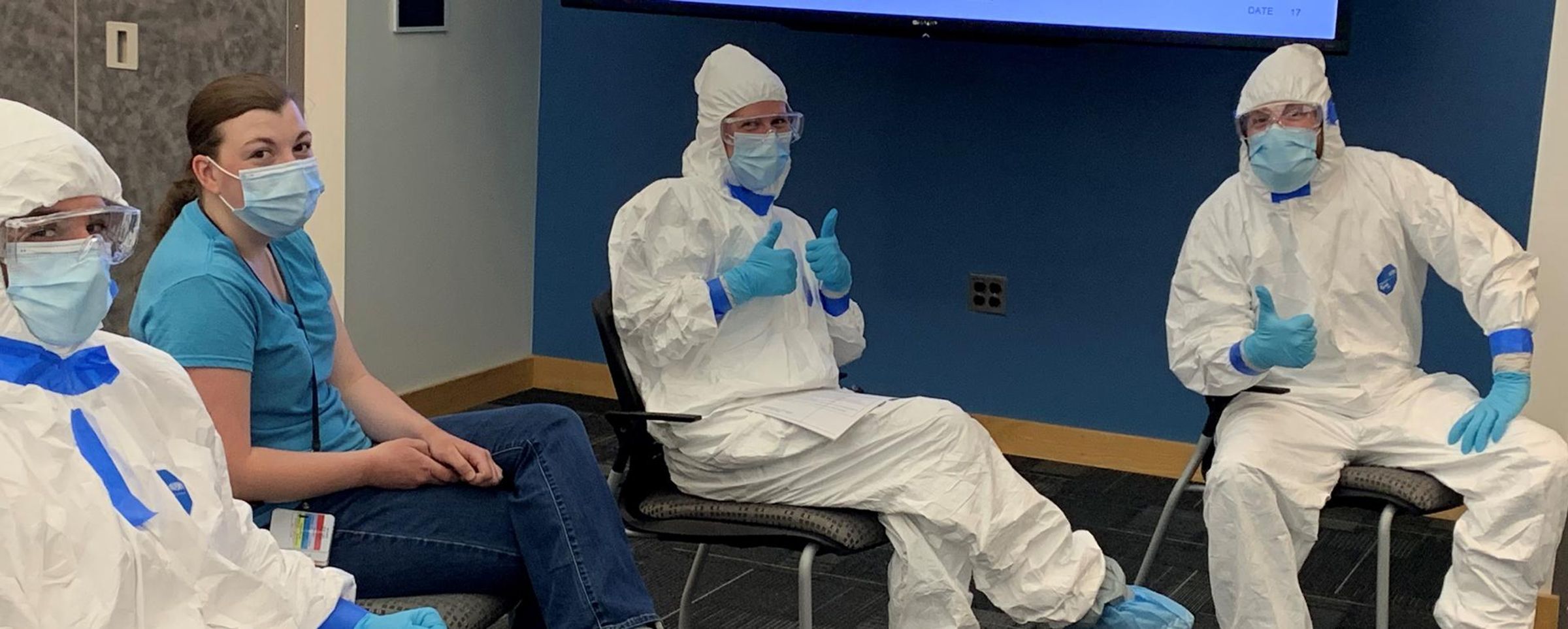 Jennifer and three of her colleagues sit in a half circle while dressed in full tyvek suits, masks and gloves. Jennifer gives a thumbs up to the camera.