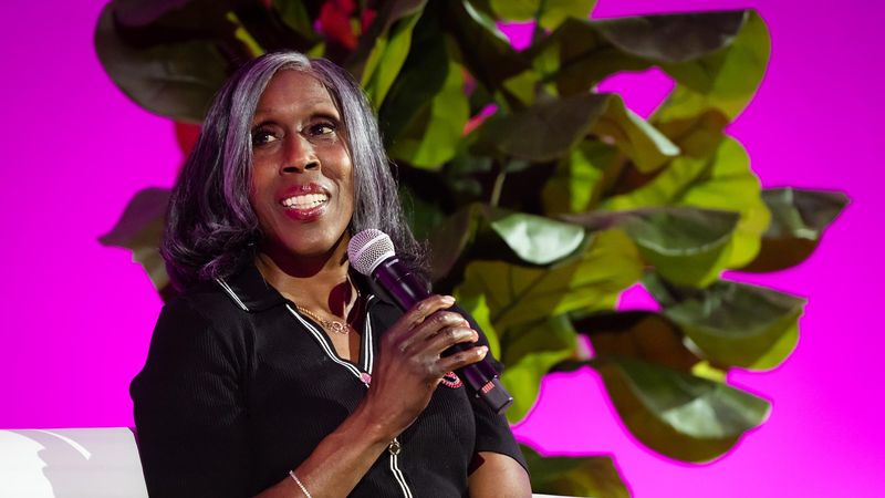 Joetta Clark Diggs takes part in the Liberty Science Center Community Evening for The Air We Share. Joetta is a smiling woman with gray hair in a black and white striped shirt, and speaks into a microphone. She sits on a white couch in front of a large green plant, with a blurred pink background.