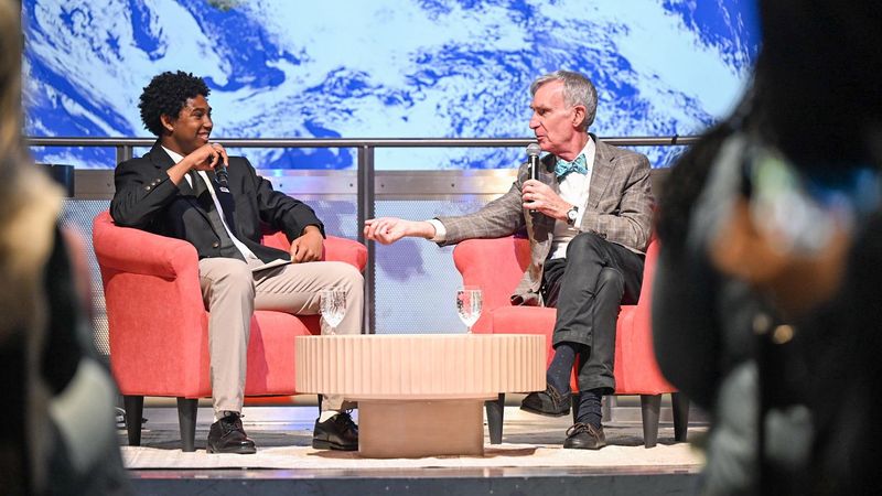 A Boys & Girls Club student sits on a stage interviewing Bill Nye the Science Guy at the Museum of Science in Boston in front of a live audience, discussing air quality and youth engagement.