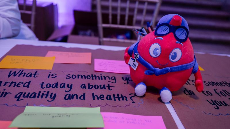 Lungston, the educational guide for The Air We Share, as a plushie with blue glasses, hat, and scarf sits on a brown cardboard display board with the handwritten prompt 'What is something you learned today about your quality and health?' Colorful Post-it notes in yellow, pink, and green are scattered around, reflecting participant responses in an interactive learning activity.