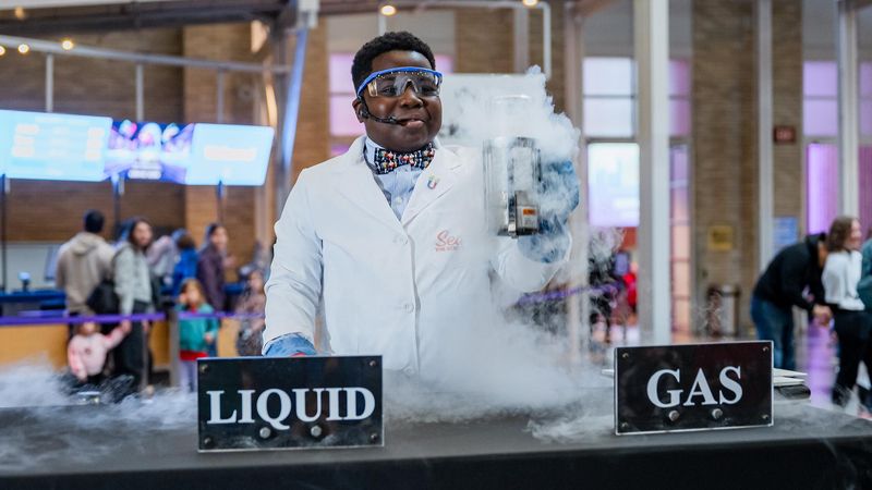 Sean the Science Kid, wearing a white lab coat, bow tie, and safety goggles, holds a steaming flask during a hands-on experiment with a Franklin Institute staff member at The Air We Share event, with table signs reading Liquid and Gas.