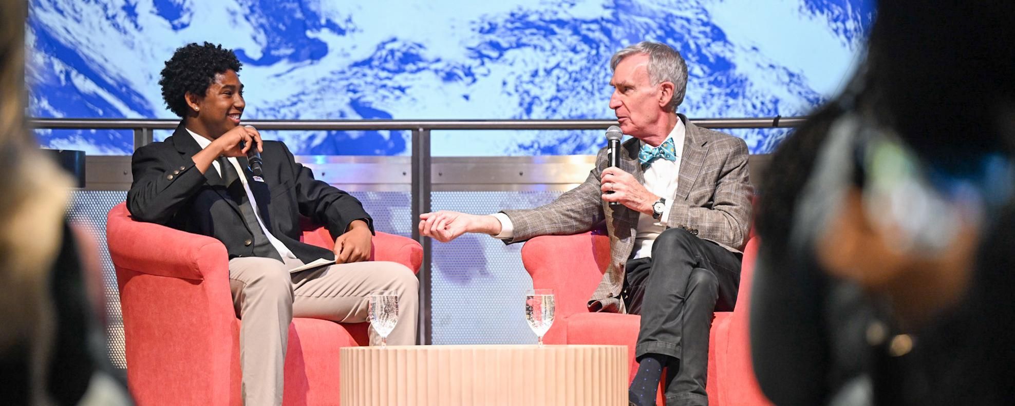A Boys & Girls Club student sits on a stage interviewing Bill Nye the Science Guy at the Museum of Science in Boston in front of a live audience, discussing air quality and youth engagement.