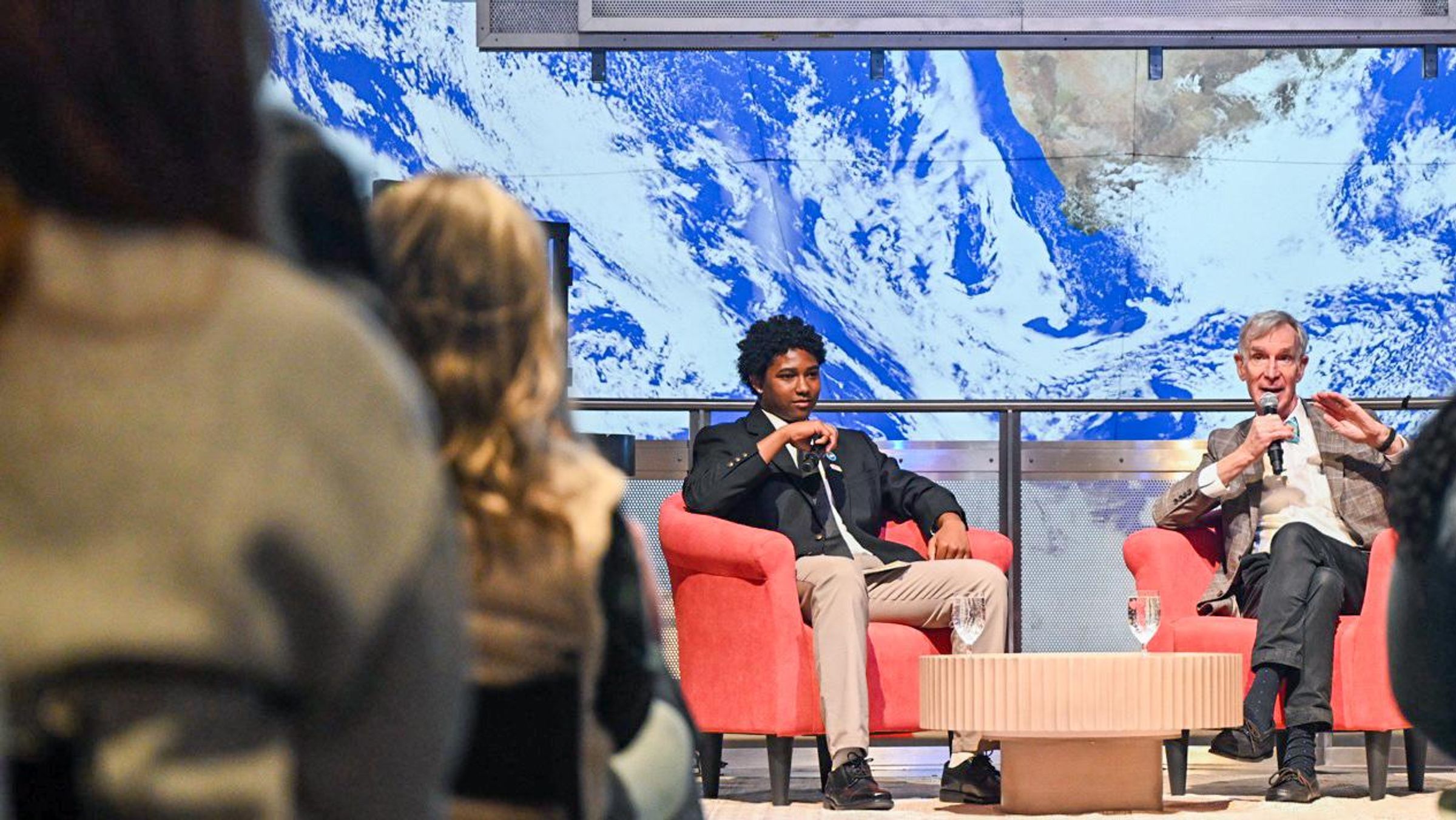 A Boys & Girls Club student sits on a stage interviewing Bill Nye the Science Guy at the Museum of Science in Boston in front of a live audience, discussing air quality and youth engagement.