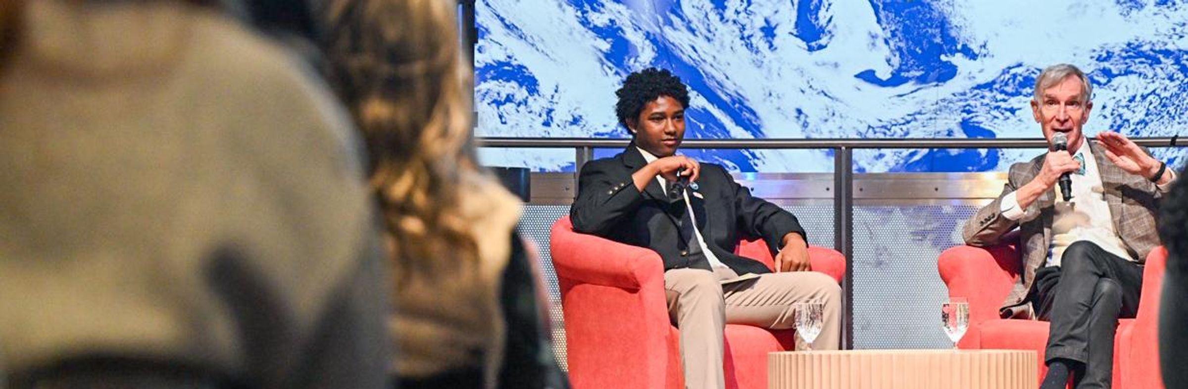 A Boys & Girls Club student sits on a stage interviewing Bill Nye the Science Guy at the Museum of Science in Boston in front of a live audience, discussing air quality and youth engagement.