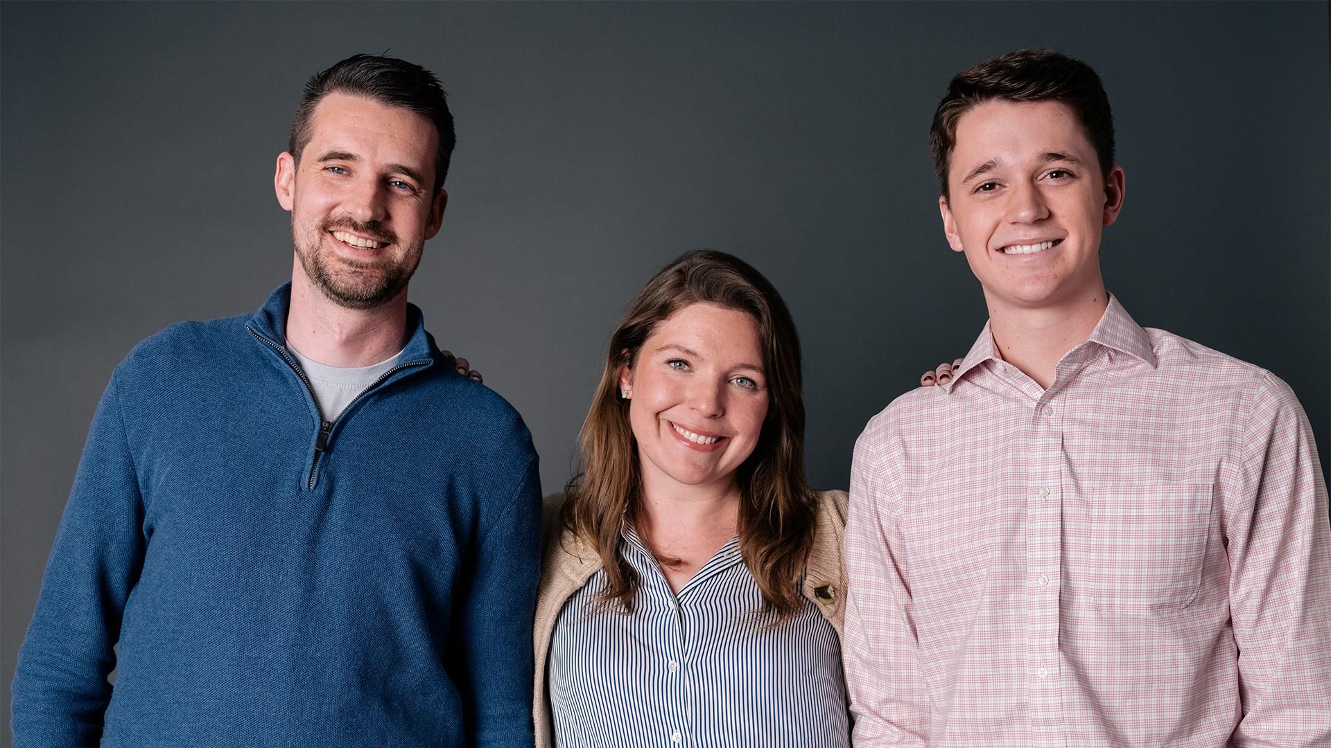 Three diabetes Sanofi Patient Ambassadors, two men and one woman, stand in a row smiling at the camera against a dark grey background.