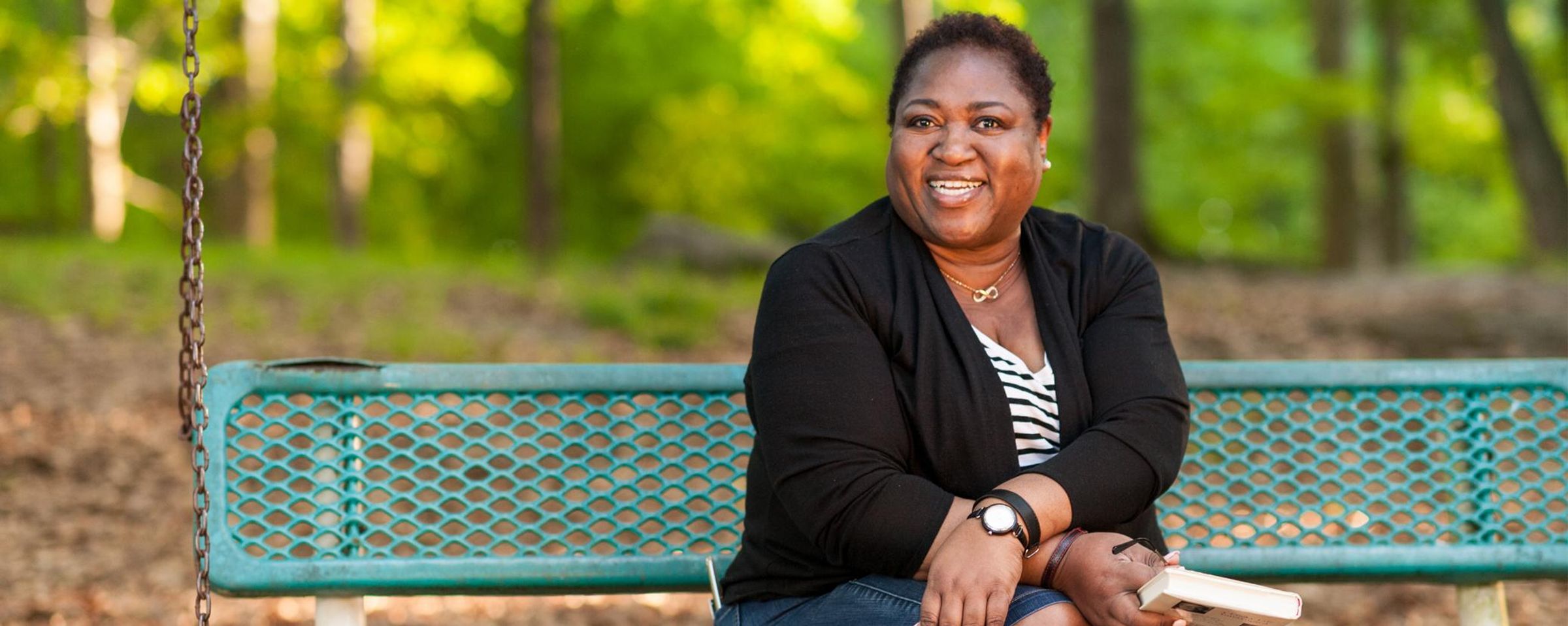 An African American woman sits on a green bench holding a book and smiling at the camera.