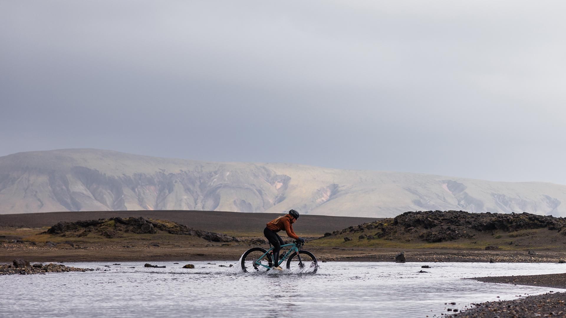 Cyclist riding a bike through a shallow body of water in the mountains of Iceland.