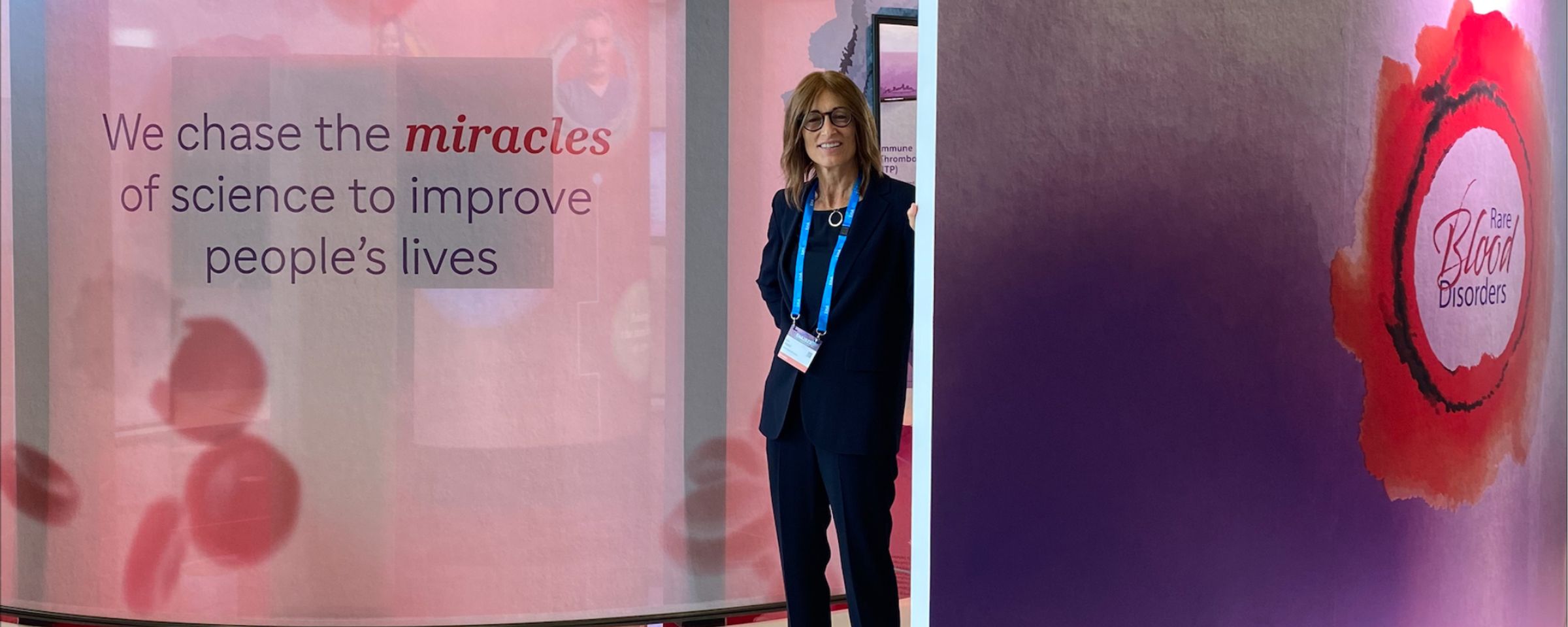 Dr. Karin Knobe, dressed in a suit with a lanyard, is standing in a medical conference booth that has images of red blood cells; on the left, there is text that says, “We chase the miracles of science to improve people’s live”; on the right, there is text that says, “Rare Blood Disorders.”