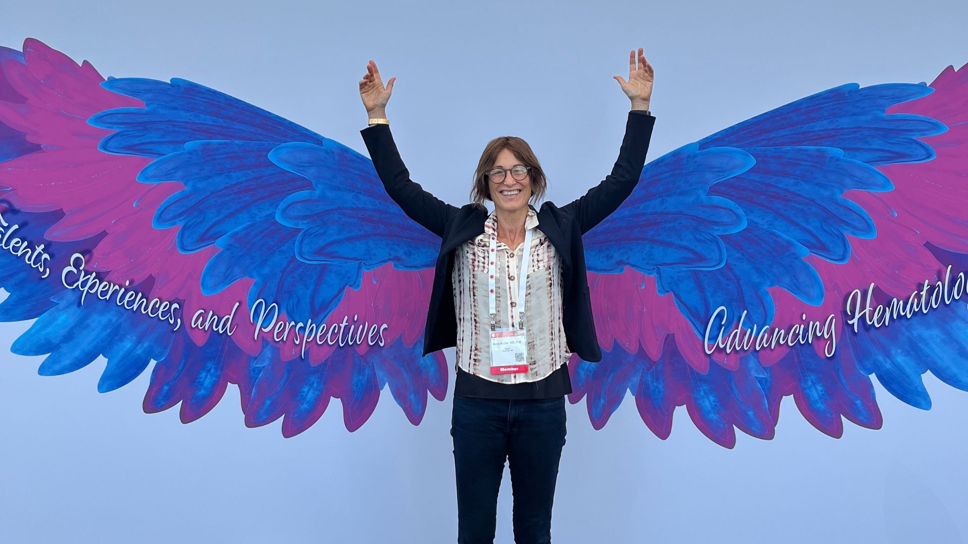 Dr. Karin Knobe, dressed in a suit with a lanyard, poses in front of an art installation. The light blue wall has a set of wings that are wider than an arm’s length in blue, purple, and pink with words written over them. Some words are cut off but what is visible says “Experiences, and Perspectives” on the left and “Advancing Hematology” on the right.