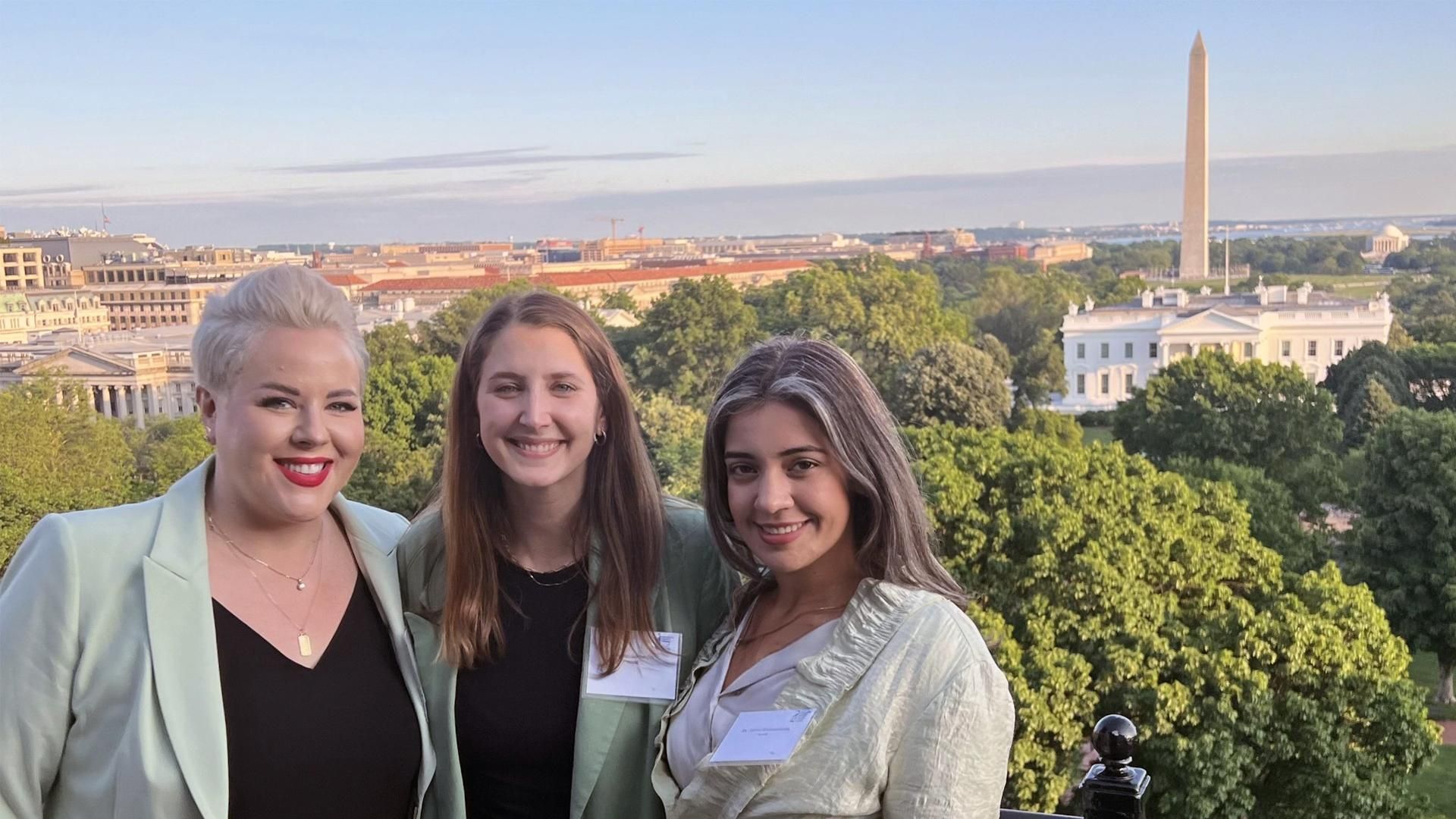 Rachel Solomon is standing between two other U.S. Public Affairs & Patient Advocacy teammates who are on a balcony with the White House and other iconic Washington D.C. buildings visible in the background.
