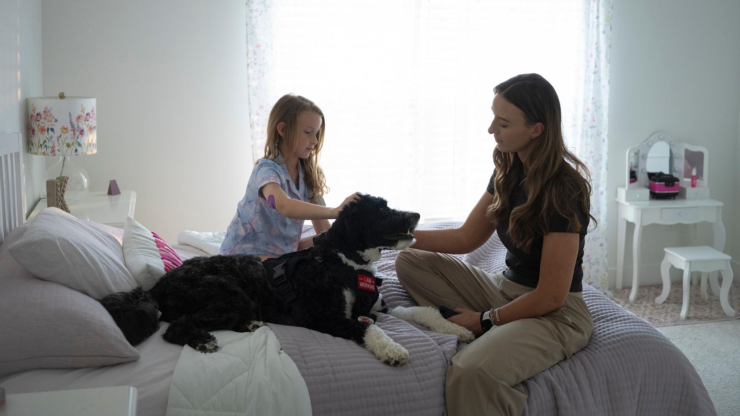 Mother and young daughter with type 1 diabetes sitting together petting their dog in a bright bed room