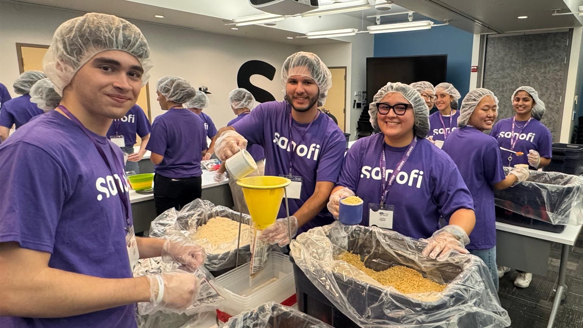 A group of young professionals are seen around several tables all wearing purple t-shirts with the Sanofi logo, as well as hair nets and plastic gloves. They are packaging rice and other foods in bags as part of an effort to help flight global food insecurity