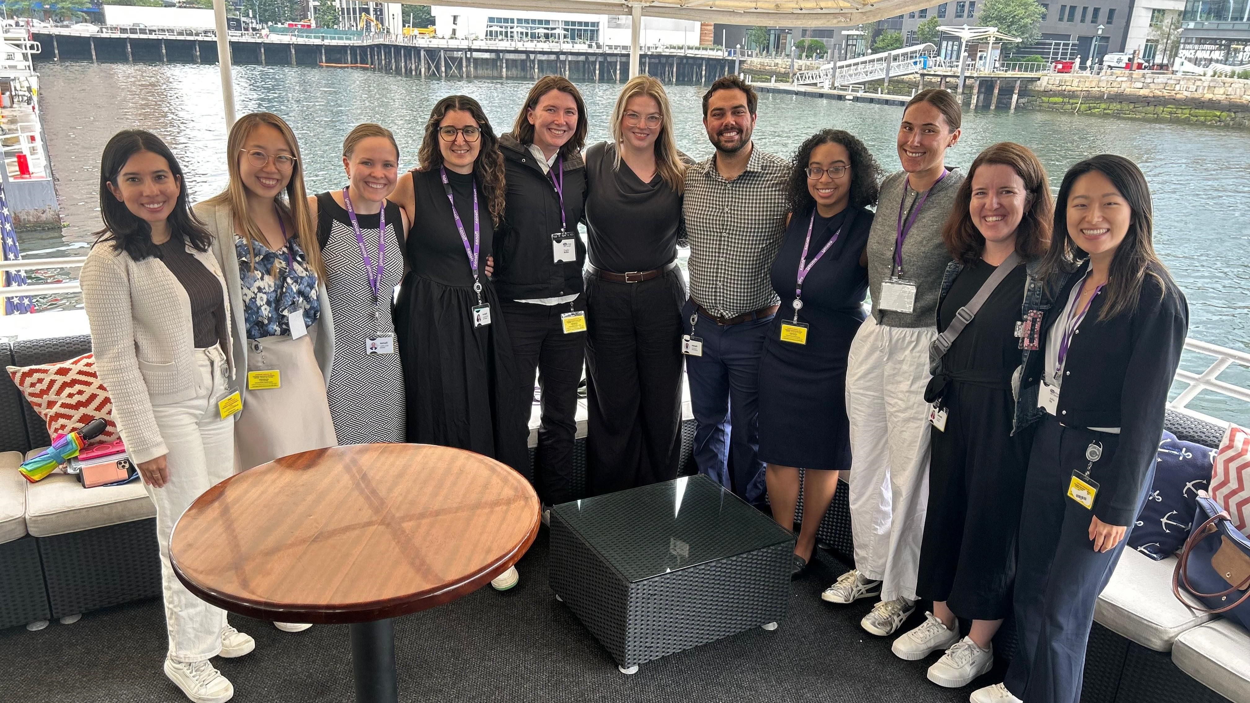 Eleven young professionals stand posing to the camera all wearing business casual attire. They are standing on a boat which is docked in a local harbor.