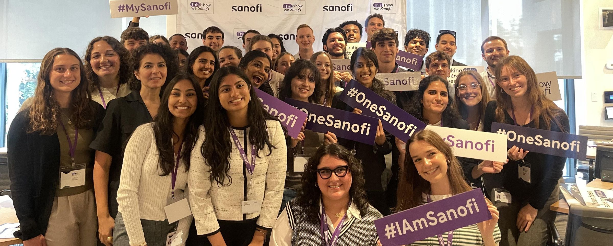 A large group of 30 to 40 interns pose, smiling at the camera in front of a Sanofi step-and-repeat. Several of them are holding signs that say “#IAmSanofi,” “#MySanofi,” or “#OneSanofi.”