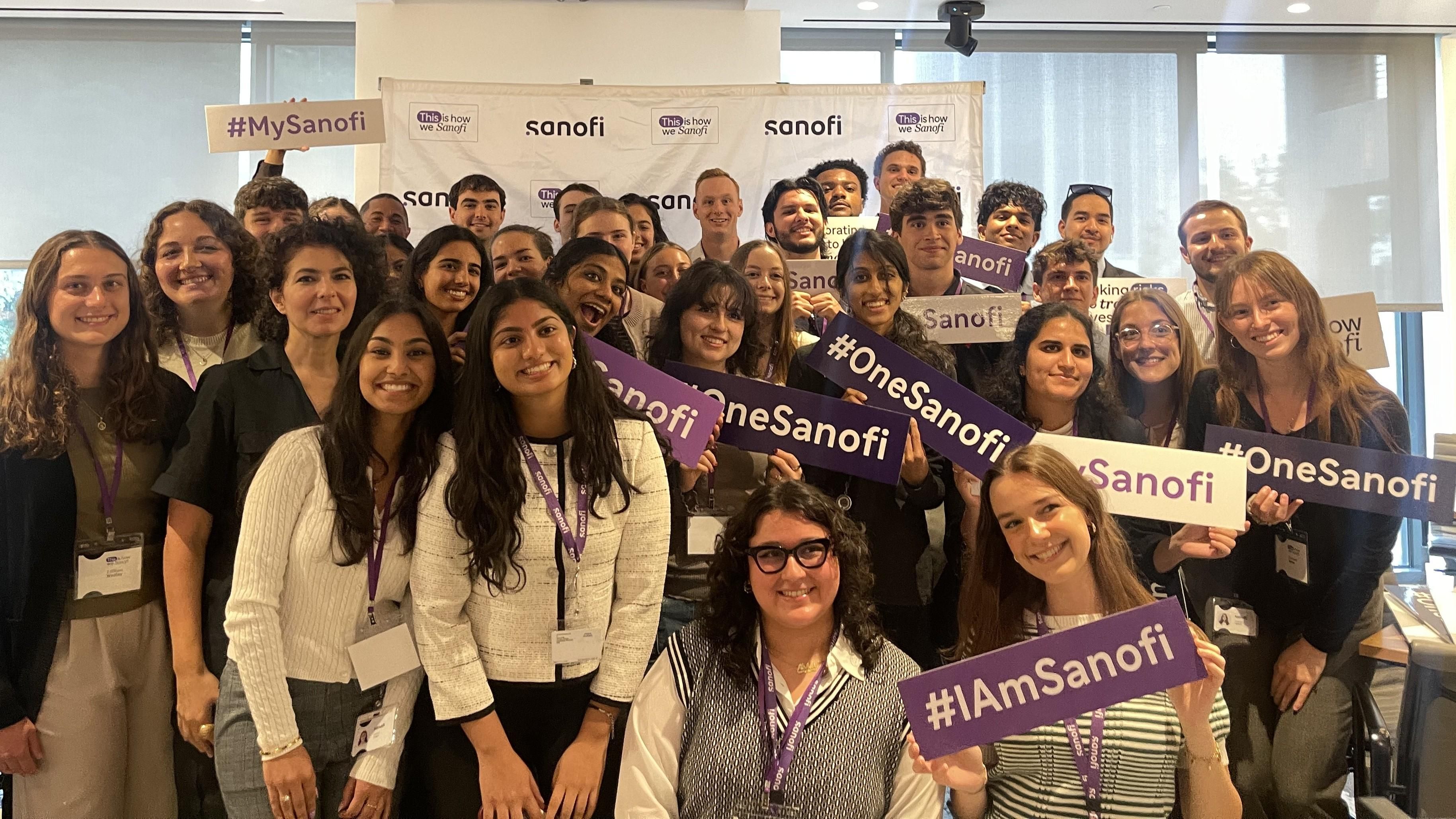 A large group of 30 to 40 interns pose, smiling at the camera in front of a Sanofi step-and-repeat. Several of them are holding signs that say “#IAmSanofi,” “#MySanofi,” or “#OneSanofi.”