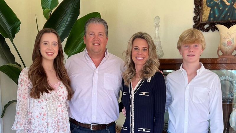 Four members of a family posing for a photo in an off-white-colored room. Family is standing in front of decorative paintings, objects, and a plant.