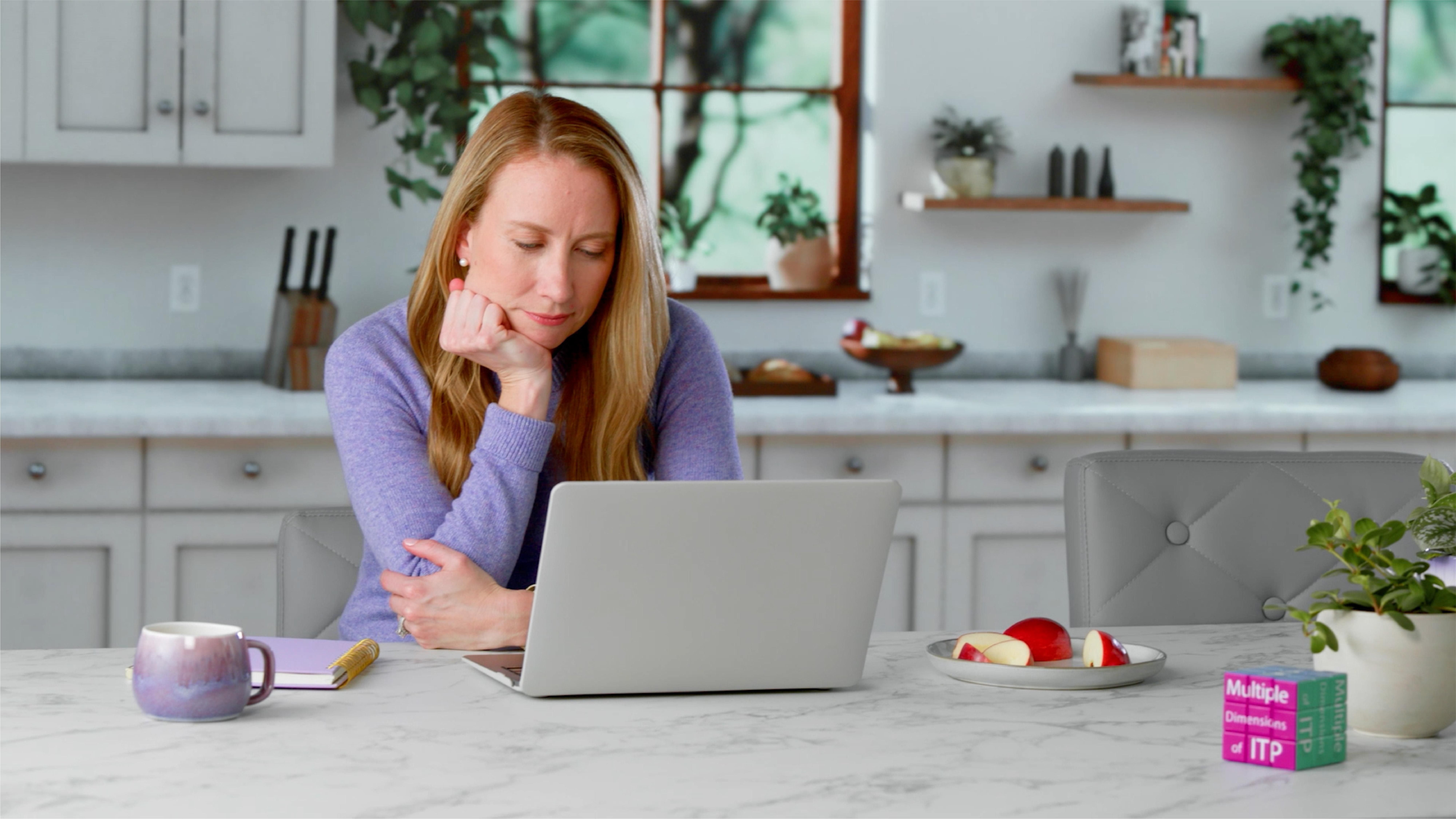 Holly, wearing a lavender sweater, sits at a white kitchen counter with a cup of tea, a cut-up red apple, and her head in her hand, looking at a laptop screen. 